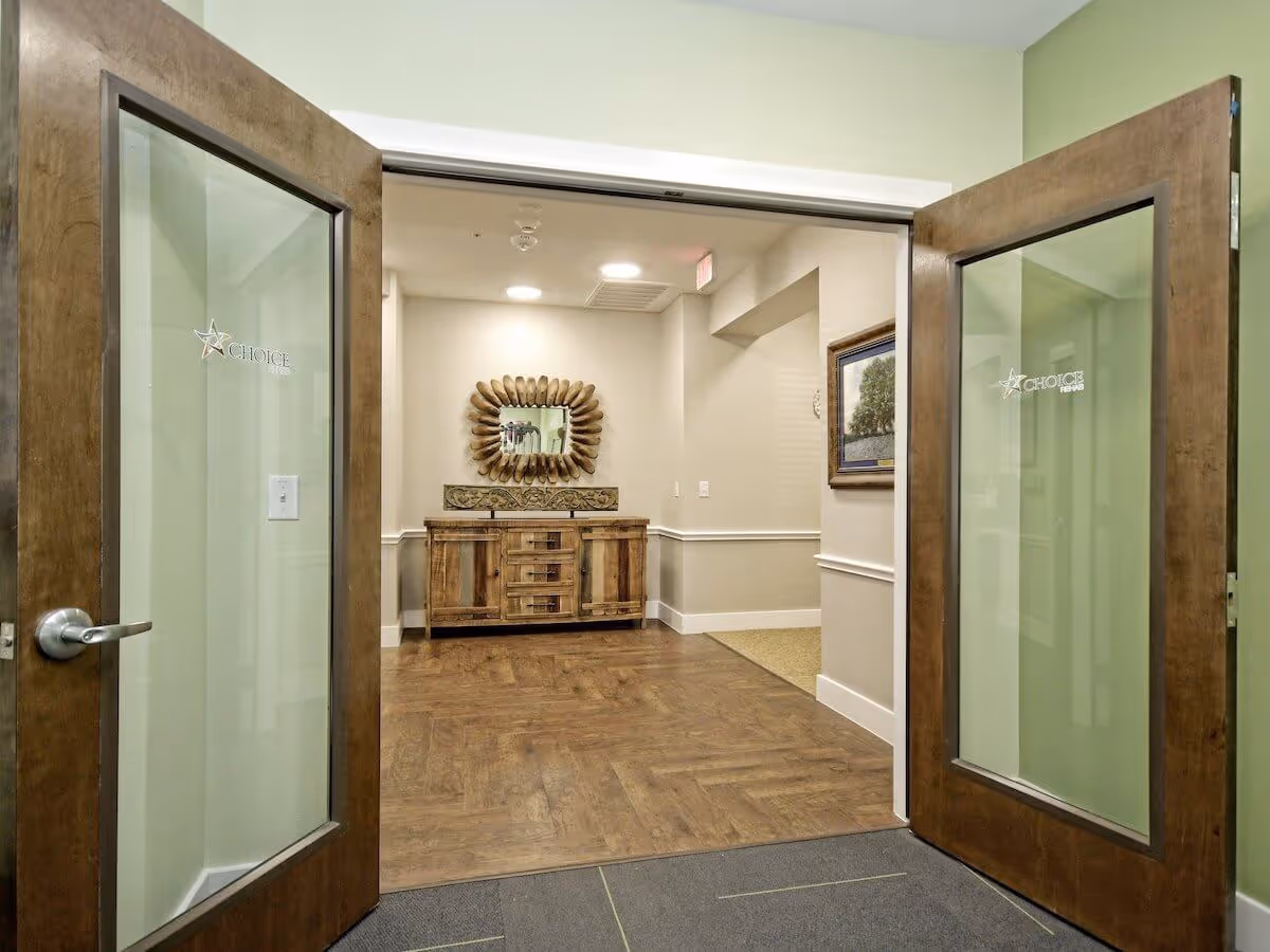 Open double wooden doors with glass panels leading into a hallway with wood flooring, a wooden sideboard with drawers and cabinets, a decorative mirror above it, and a framed picture on the right wall.