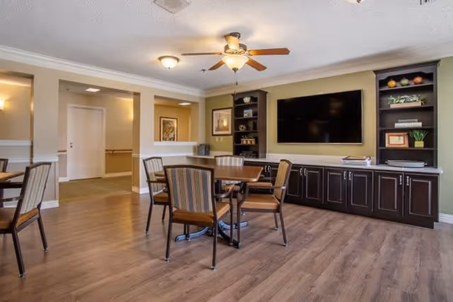 A bright common dining area with round tables and chairs, a wall-mounted TV above built-in dark cabinets, wood flooring and a ceiling fan.