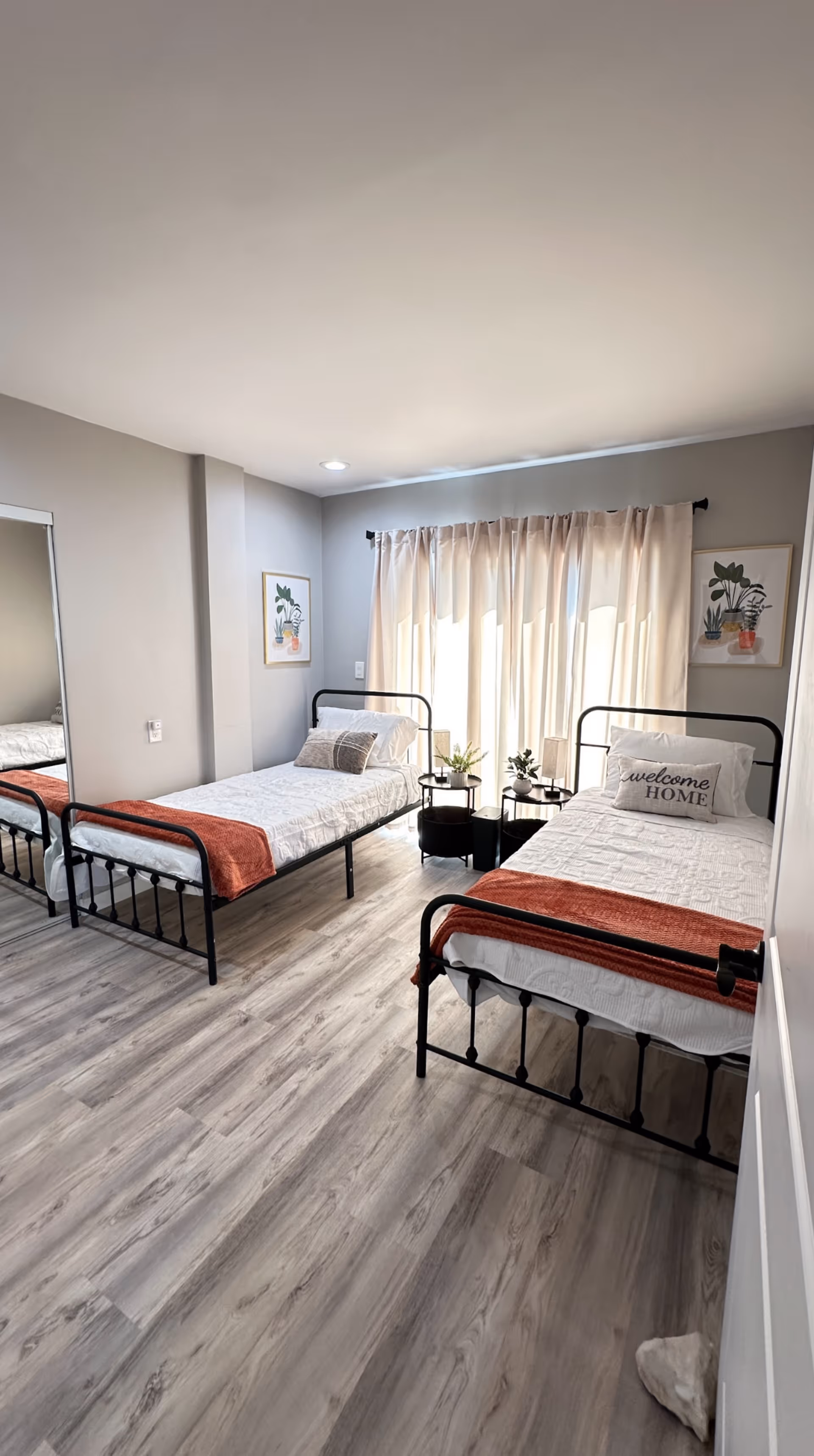 A bright bedroom with two single metal frame beds, each with white bedding and a rust-colored throw blanket at the foot. One bed has a pillow with the text 'welcome HOME'. Between the beds are two small round black tables with potted plants. The room has light wood flooring, light gray walls, a large window with sheer beige curtains, and two framed botanical prints on the walls.