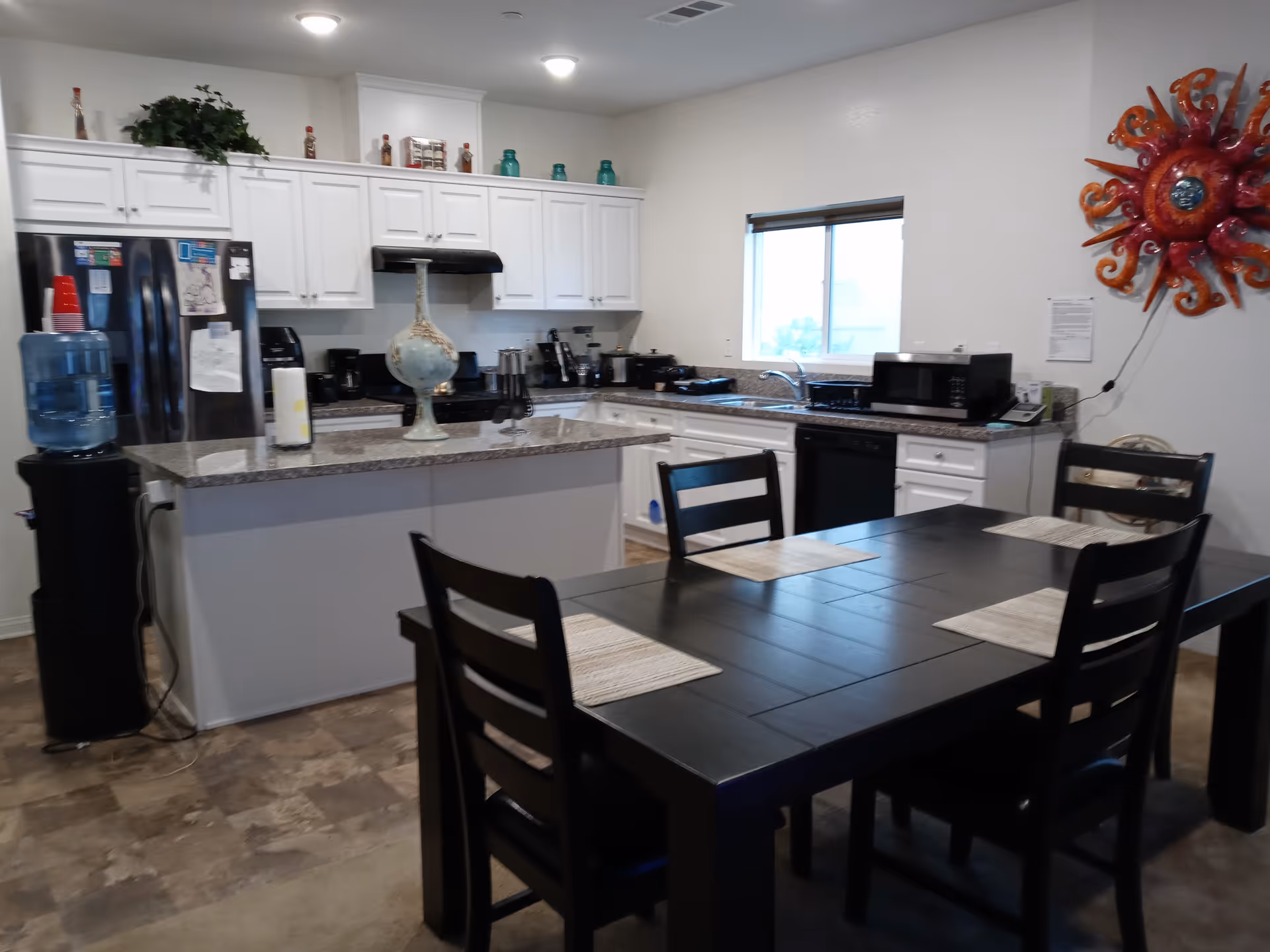 A kitchen and dining area with white cabinets, a granite countertop island, and a black dining table with four chairs. The kitchen includes a refrigerator, microwave, dishwasher, coffee maker, and a water dispenser. A colorful sun-shaped wall decoration is mounted on the wall near a window.