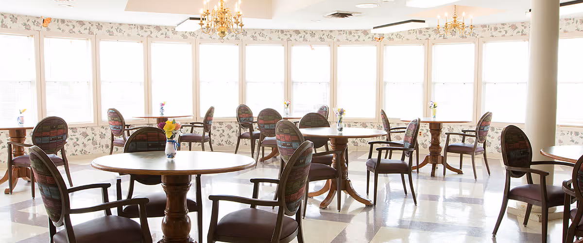 A bright dining room with multiple round wooden tables and cushioned chairs arranged neatly. Each table has a small vase with colorful flowers. Large windows with floral wallpaper border let in ample natural light, and chandeliers hang from the ceiling.