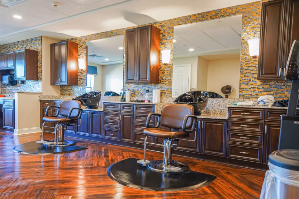 Interior view of a salon area with two brown salon chairs in front of large mirrors mounted on a wall with mosaic tile backsplash. The room features dark wood cabinetry, granite countertops, and wooden flooring.