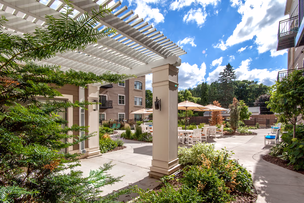 Outdoor courtyard area at The Waters of Highland Park featuring a white pergola, patio tables with umbrellas, chairs, and lush green landscaping under a partly cloudy blue sky.