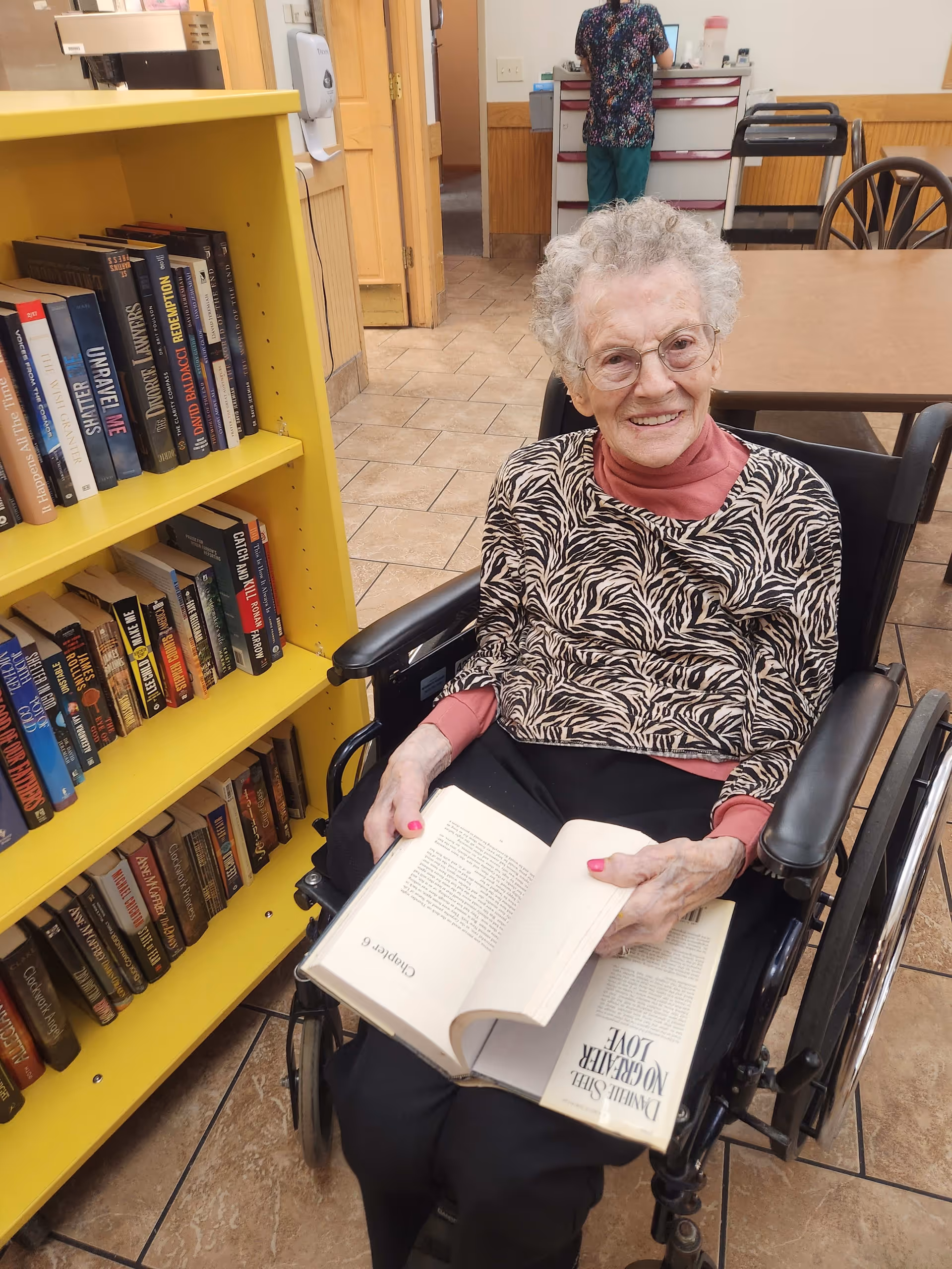 An elderly woman with curly gray hair and glasses sits in a wheelchair, smiling and holding an open book. She is wearing a zebra-patterned top over a pink long-sleeve shirt. To her left is a yellow bookshelf filled with books. In the background, a person in scrubs stands near a counter in a room with tiled floors and wooden furniture.