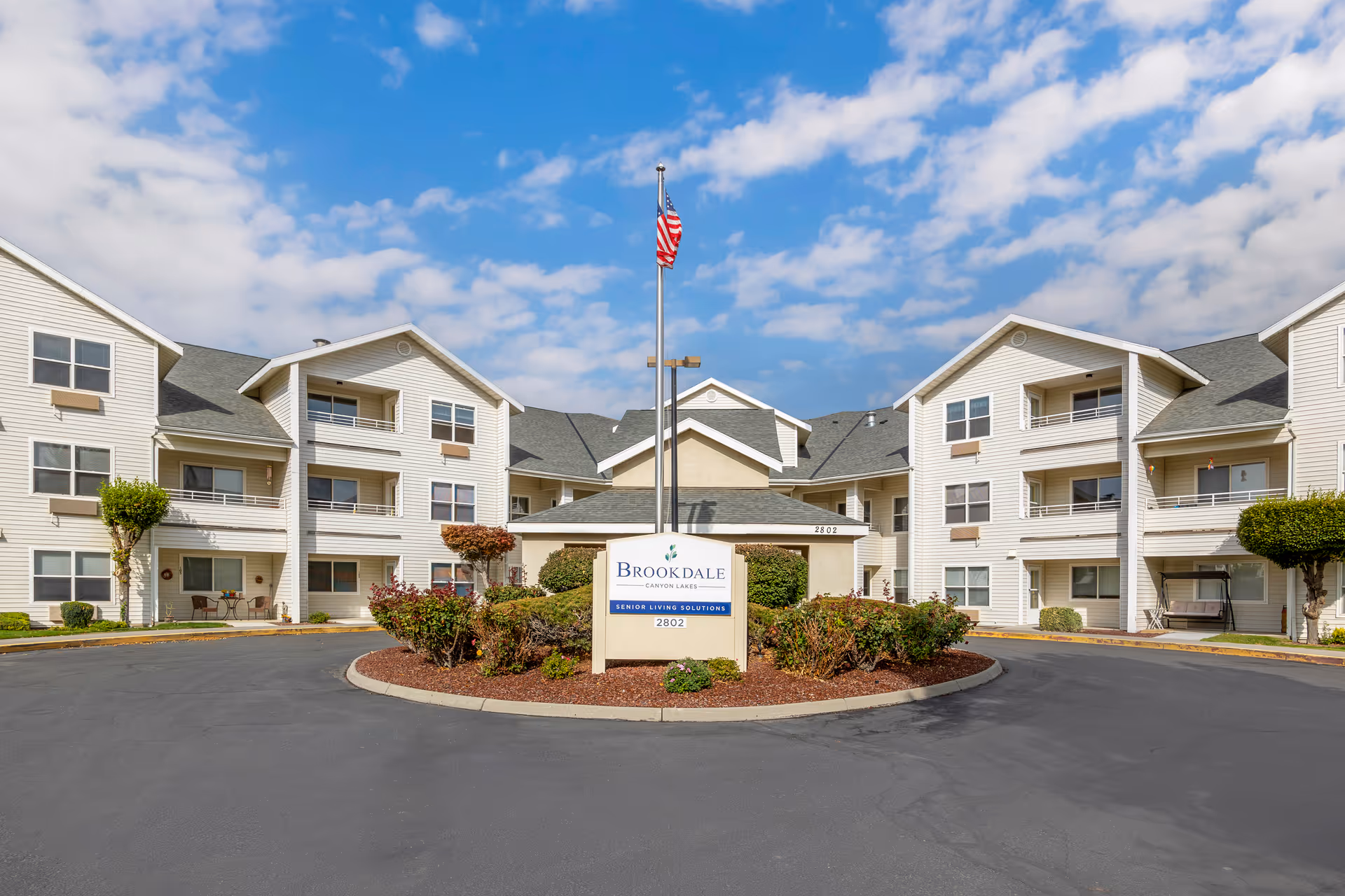 Front view of a three-story Brookdale Canyon Lakes senior living building with a central sign, circular driveway, landscaping, and an American flag.
