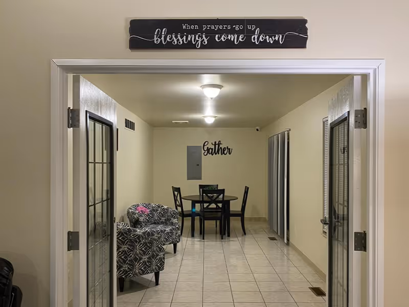 View through double glass doors into a room with tiled floor, two patterned armchairs on the left, and a round dining table with four chairs at the far end. Wall decor includes a sign above the door reading 'When prayers go up blessings come down' and another sign on the far wall that says 'Gather'.