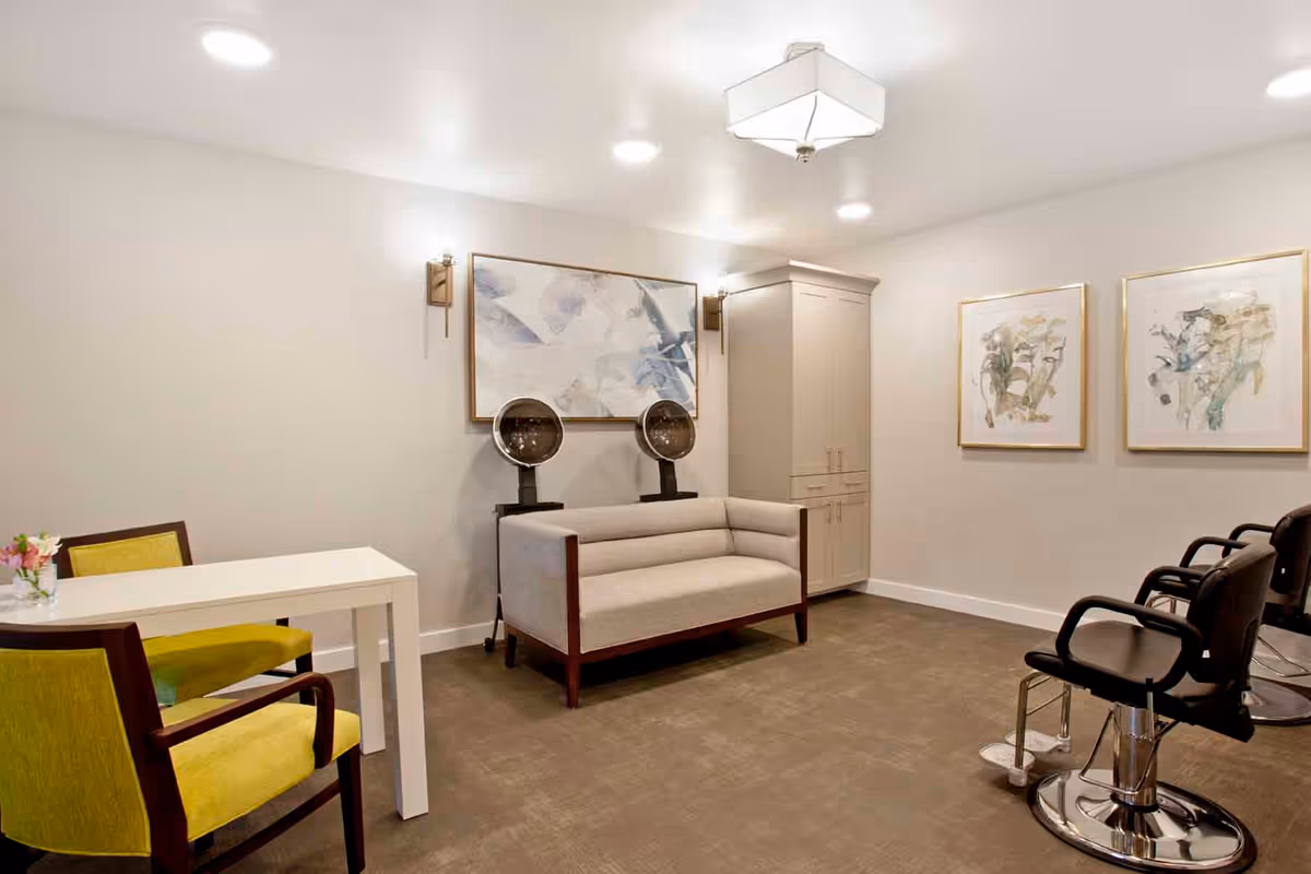 Small salon-style interior with hooded hair dryers, salon chairs, a small sofa, table and wall art.