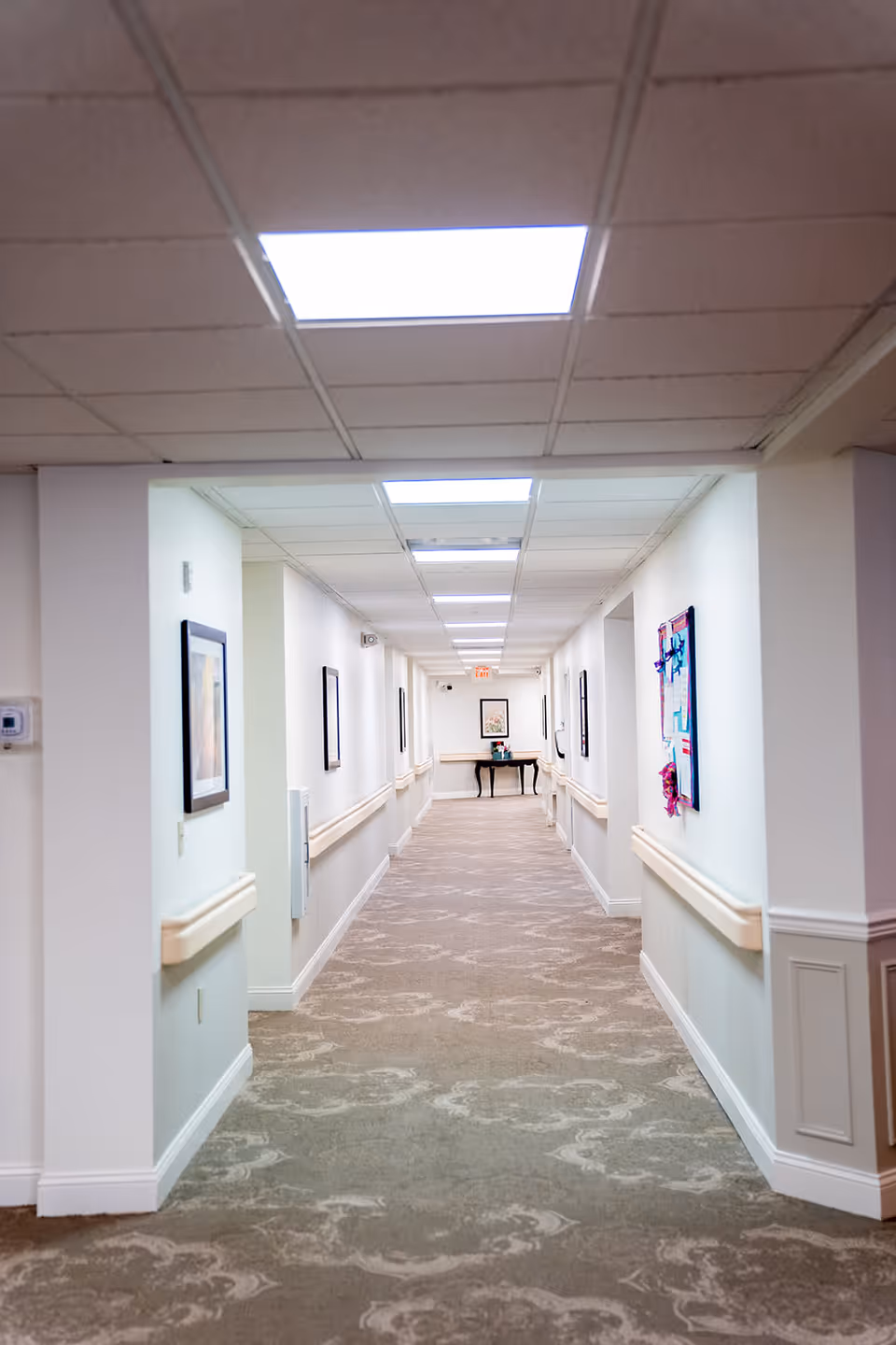 A long, well-lit hallway in a senior living facility with patterned carpet, white walls, handrails on both sides, framed artwork on the walls, and a small table with a flower arrangement at the far end.