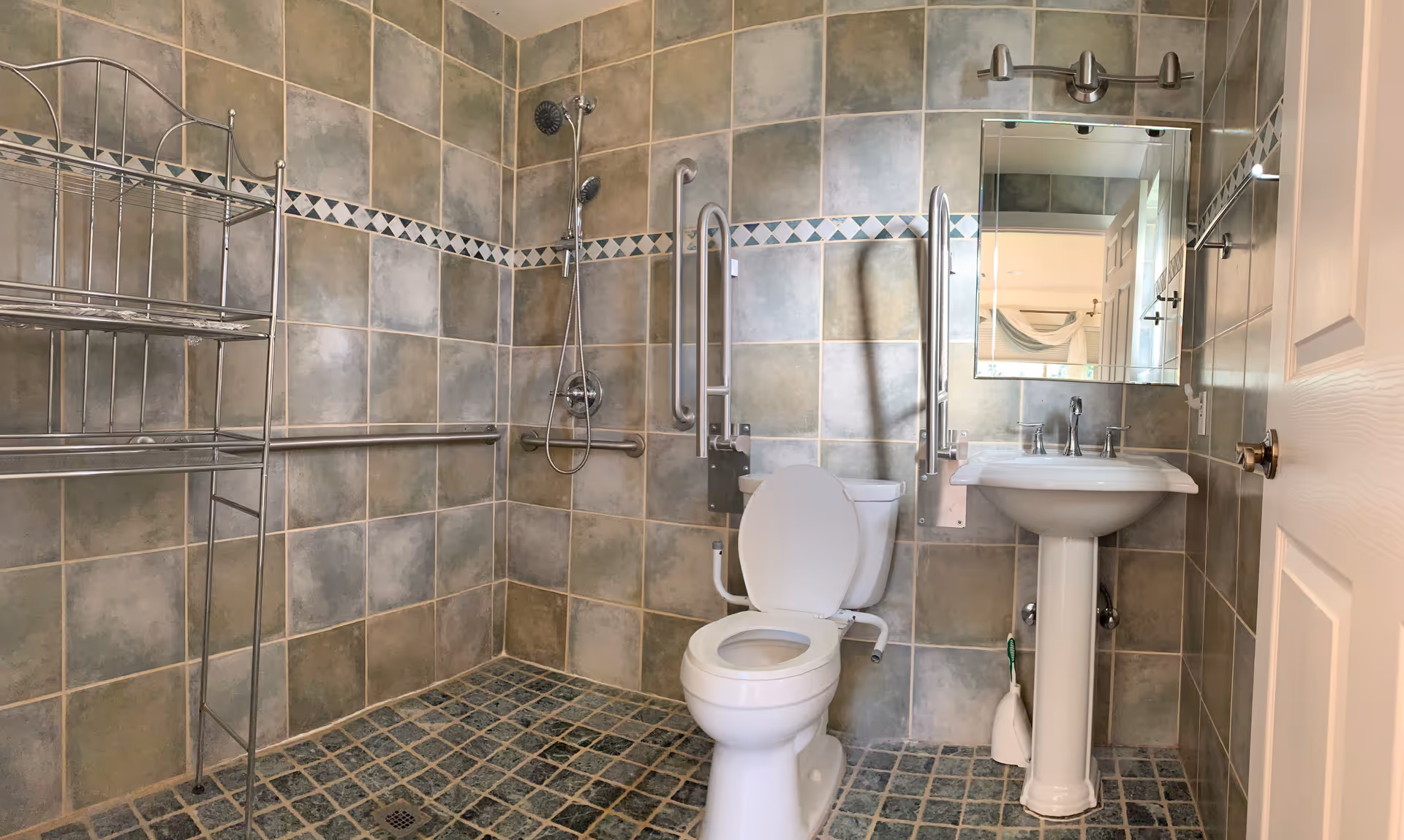 A tiled bathroom with a toilet, pedestal sink, and a walk-in shower area. The walls and floor are covered with square tiles in shades of green and beige. There are metal grab bars around the toilet and shower for accessibility, a metal rack for storage, and a mirror above the sink. The bathroom door is partially open.