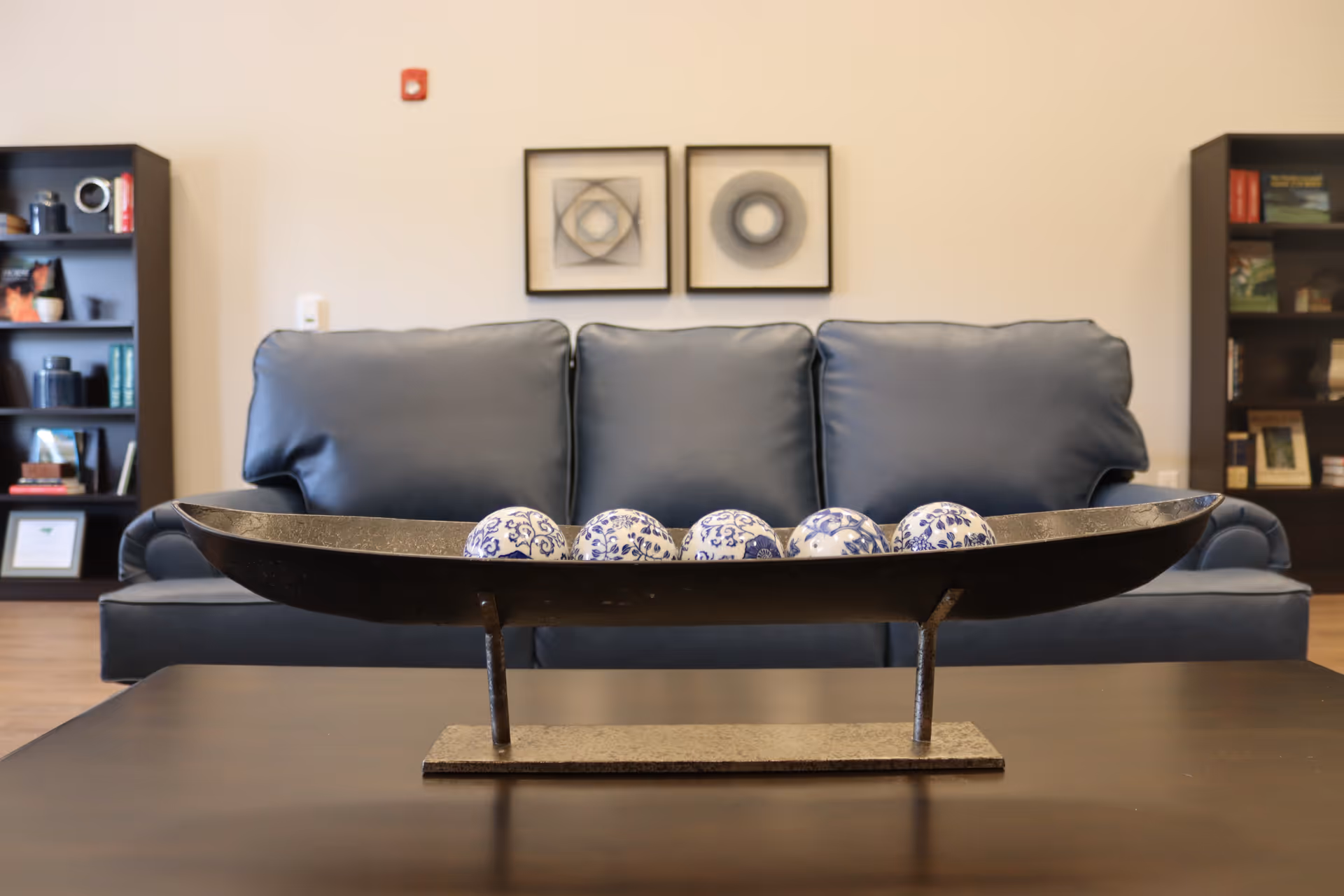 A modern living room with a dark blue leather sofa in the background and a decorative metal tray with five blue and white patterned ceramic balls on a dark wooden coffee table in the foreground. Two framed abstract artworks hang on the beige wall behind the sofa, and there are two dark wooden bookshelves filled with books and decorative items on either side of the sofa.