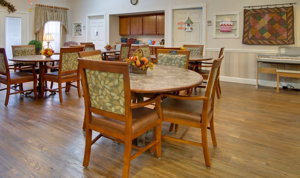 A well-lit dining room with round tables and wooden chairs featuring floral patterned cushions. The room has wooden flooring, a piano against the wall, decorative wall hangings, and a clock above a set of cabinets. There are windows with curtains and a lamp on a side table in the background.