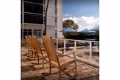 Wooden lounge chairs on a paved patio beside a pool with blue umbrellas and a multi-story building in the background under a partly cloudy sky.
