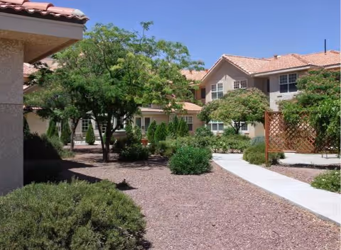 Courtyard with a paved walkway, trees and shrubs in front of two-story beige buildings with red tile roofs under a clear blue sky.