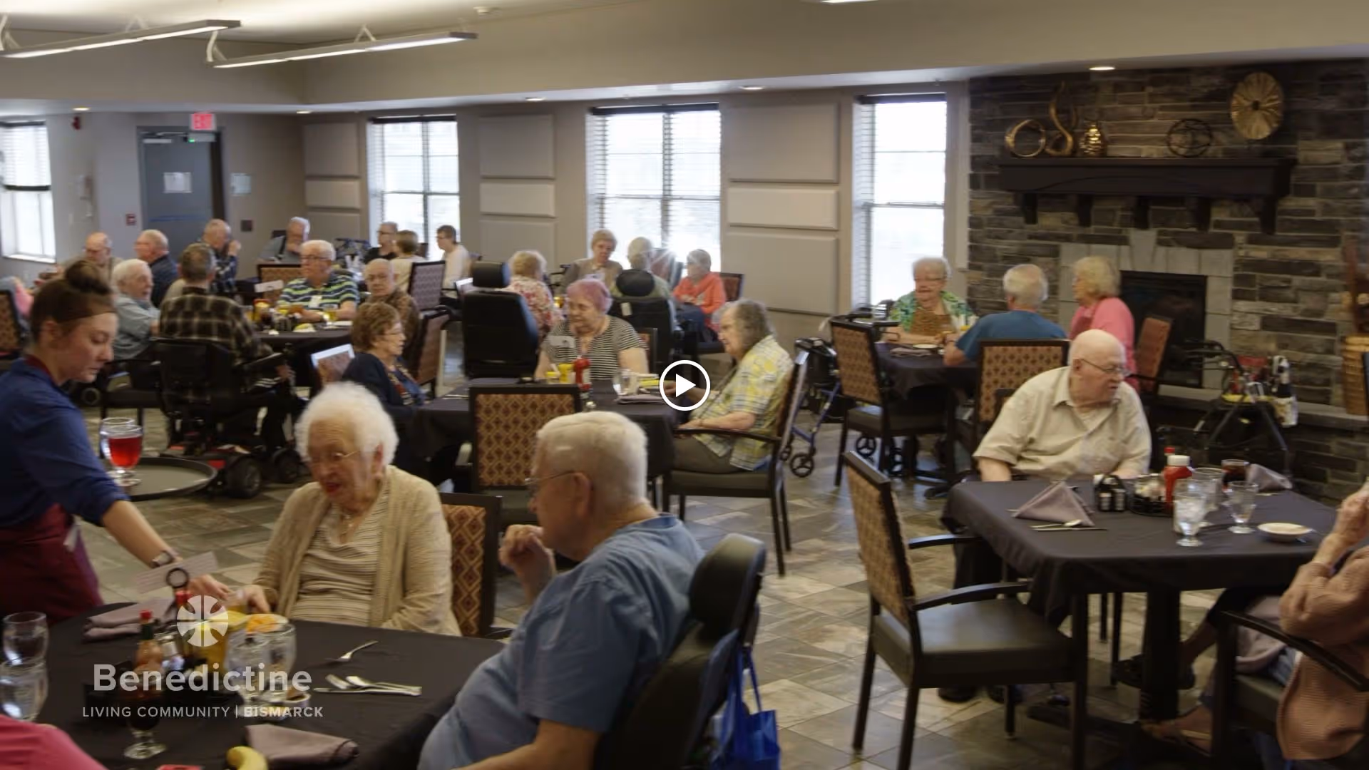 A dining room in a senior living community with elderly residents seated at multiple tables, some in wheelchairs, enjoying a meal. A staff member is serving food to one of the residents. The room has large windows, a stone fireplace, and decorative items on the mantel.