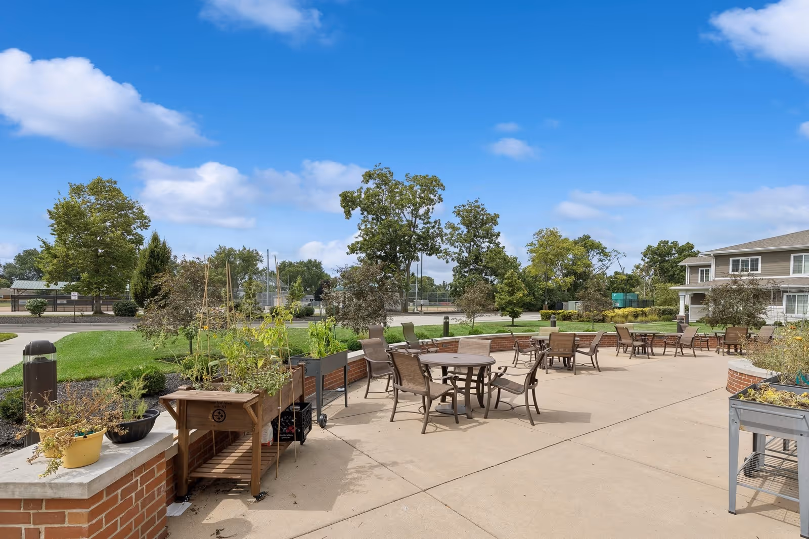 Outdoor patio area with round tables and chairs, planters, and landscaped lawns beside a two-story building.