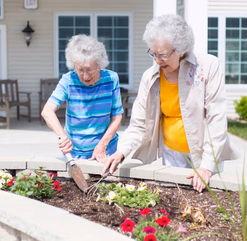Two elderly women gardening together outside, tending to a flower bed with red and white flowers in front of a building with large windows and a porch area with chairs.