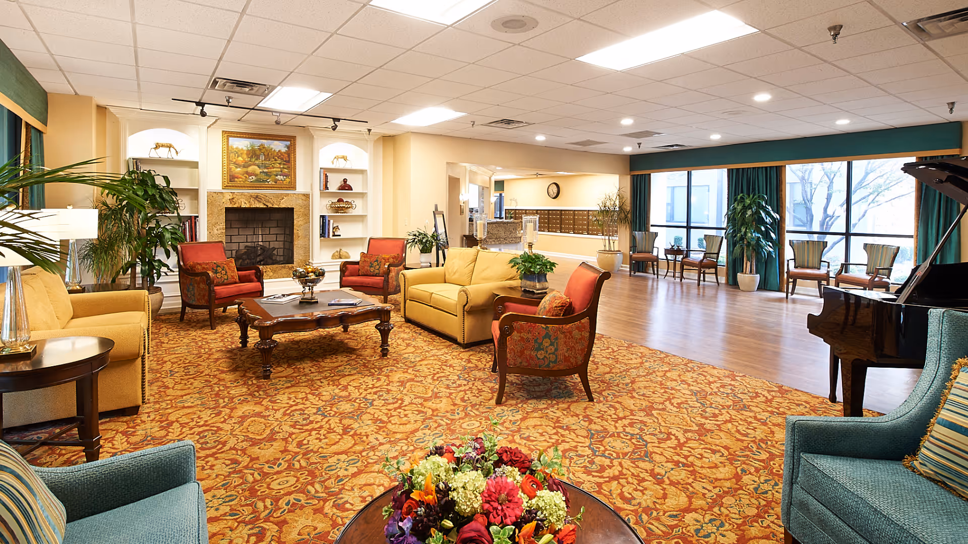 A spacious and well-lit senior living facility common area with comfortable seating including armchairs and sofas arranged around a wooden coffee table on a patterned carpet. There is a fireplace with built-in shelves, large windows with green curtains, several potted plants, and a grand piano on the right side of the room.
