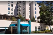 Front exterior of a multi-story senior living building with a teal awning over the main entrance and surrounding trees.