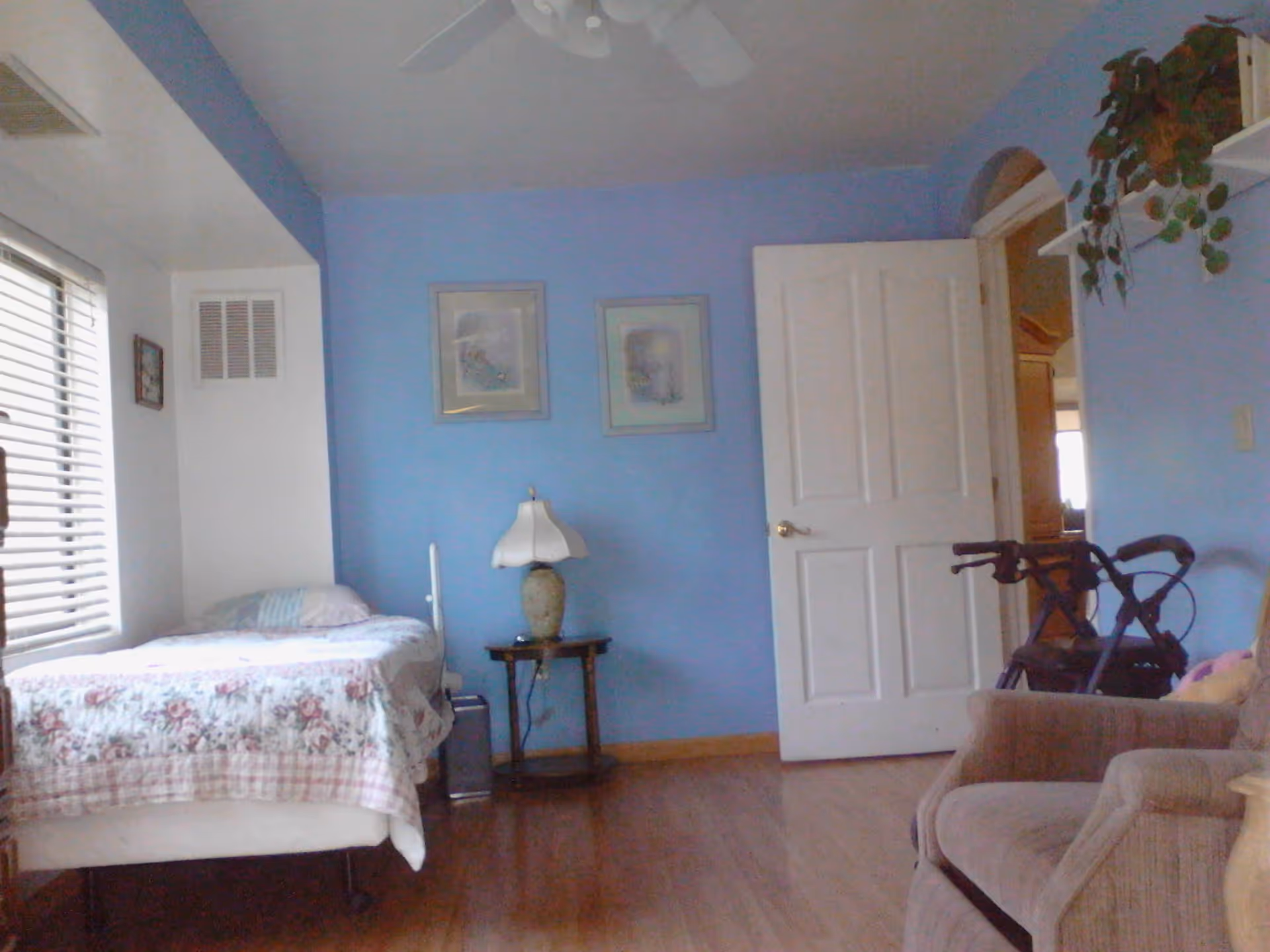A bedroom with light blue walls and wooden flooring. There is a single bed with floral bedding positioned near a window with blinds. A small wooden side table with a decorative lamp is next to the bed. Two framed pictures hang on the wall above the table. An open white door leads to another room, and a brown armchair is partially visible on the right side. A walker is placed near the doorway, and a plant is on a white shelf above the armchair.