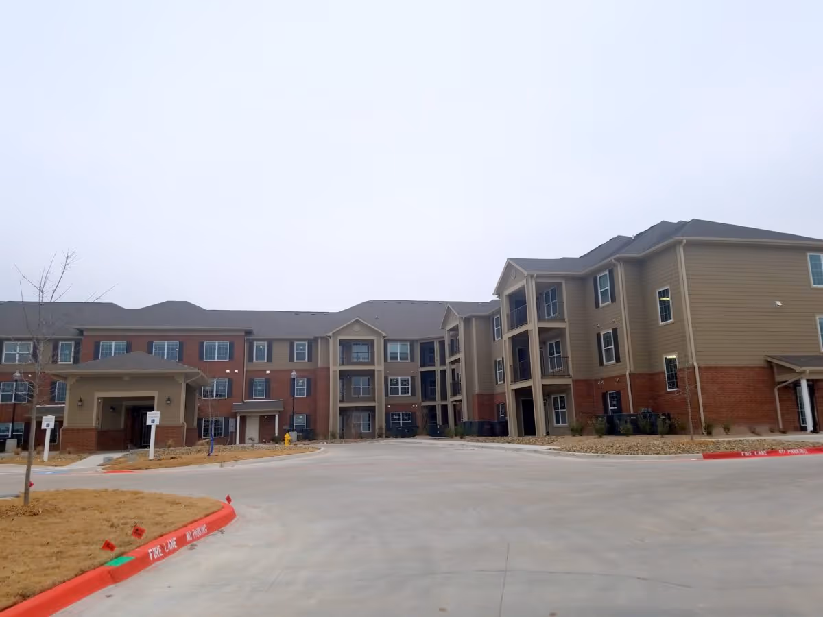 Three-story brick-and-siding senior living building with a covered main entrance and a wide driveway under an overcast sky.