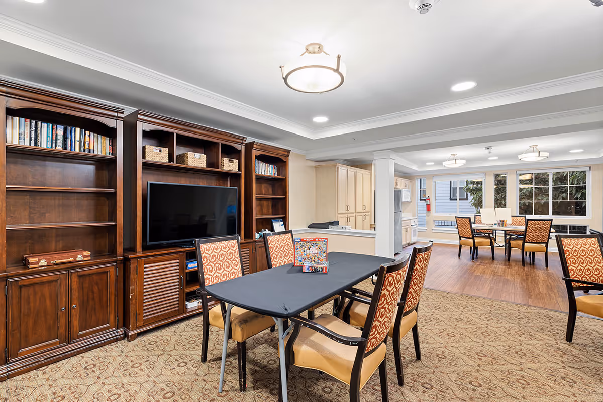 Well-lit communal living room with a large wooden entertainment wall and TV, tables and patterned chairs facing a kitchen and dining area.