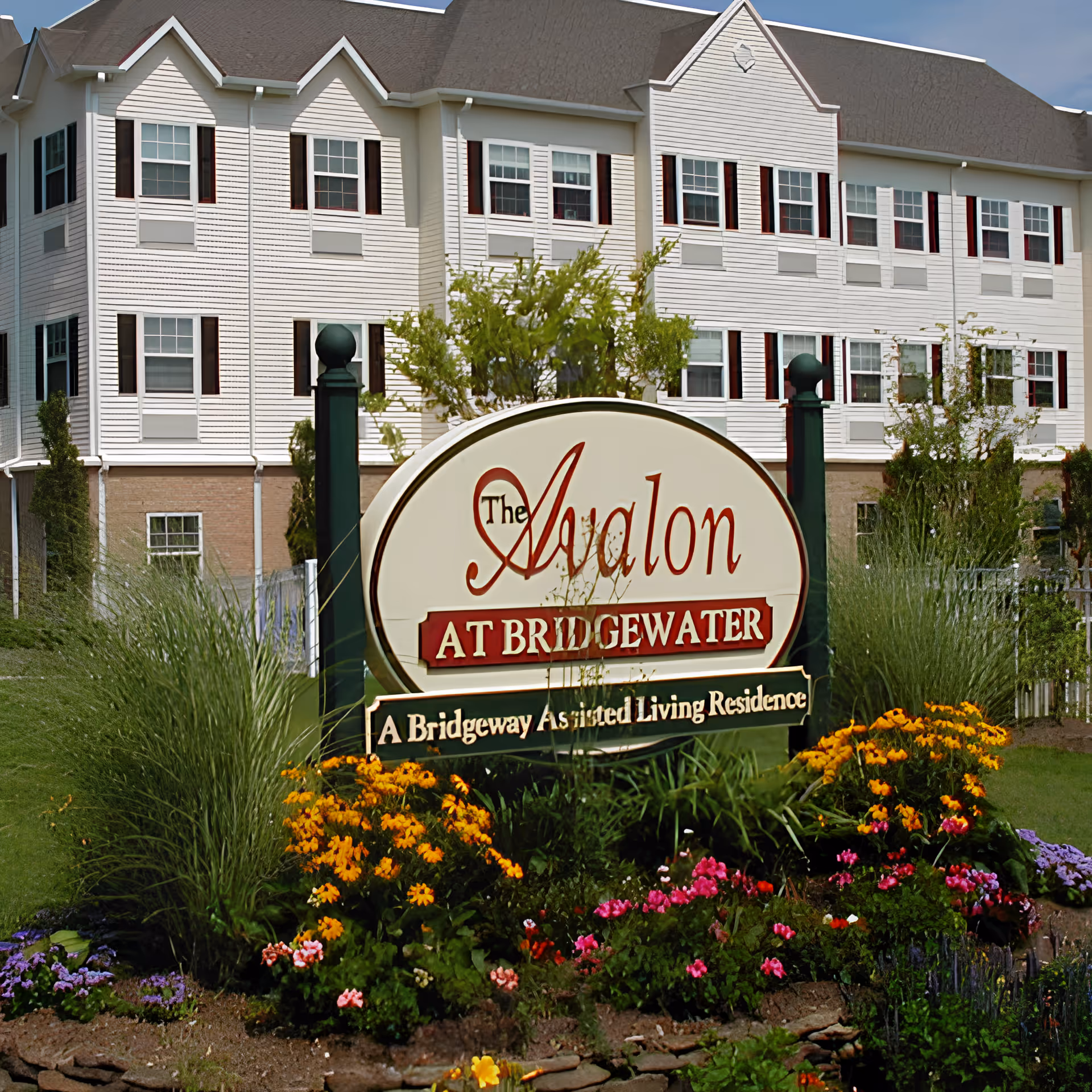 The exterior of Avalon Assisted Living at Bridgewater, showing a large white multi-story building with many windows and a dark roof. In front of the building is a landscaped garden with colorful flowers and greenery surrounding a large sign that reads 'The Avalon at Bridgewater, A Bridgeway Assisted Living Residence.'
