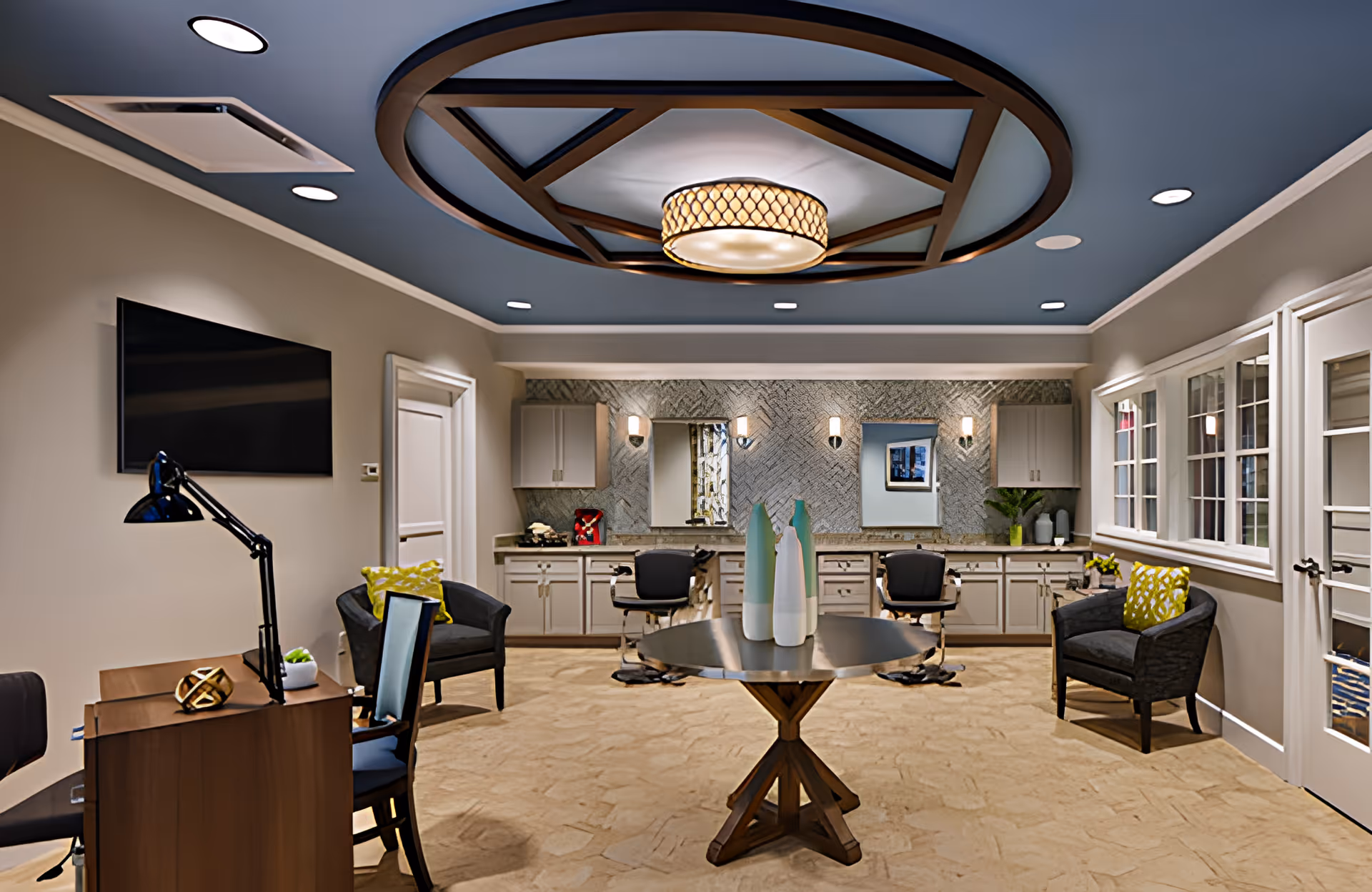 A well-lit interior room with a round wooden table in the center holding decorative vases. The room features a blue ceiling with a geometric wooden design and a circular light fixture. There are two black chairs with yellow patterned cushions on either side of the room, a desk with a lamp and chair on the left, and a wall-mounted TV. The back wall has cabinets, mirrors, and wall sconces, with a textured backsplash. Large windows and a glass door are on the right side.