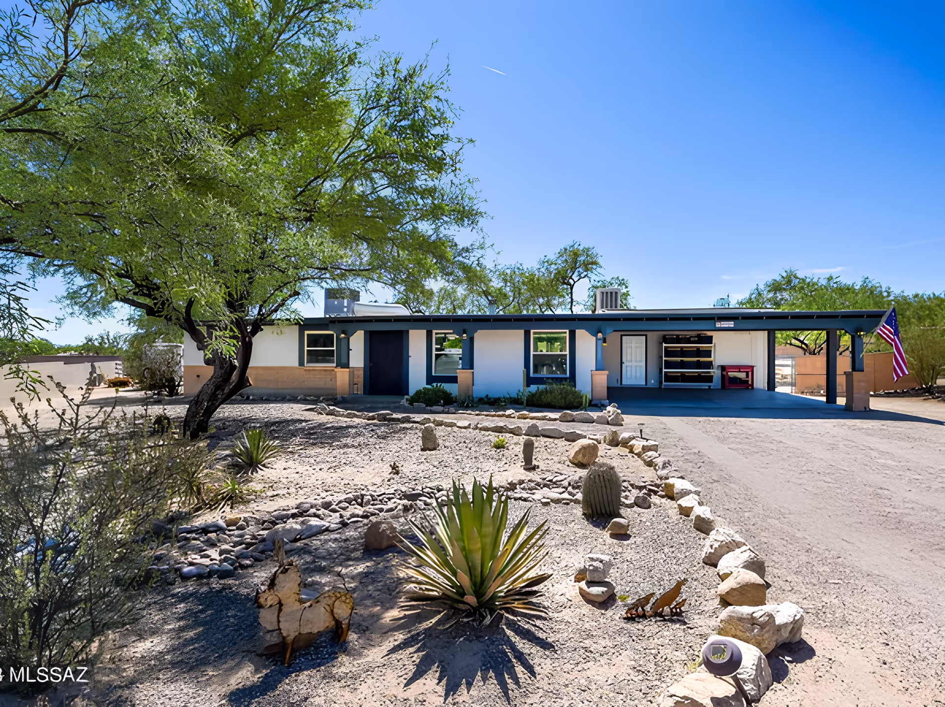Single-story ranch-style building with a covered carport, desert landscaping and an American flag under a clear blue sky.