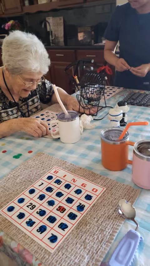 An elderly woman with white hair and glasses is playing bingo at a table covered with a blue and white checkered tablecloth. She is marking her bingo card with a pen. A bingo cage with numbered balls is on the table, along with several colorful cups with straws. Another person, partially visible, is standing nearby assisting with the game. The setting appears to be a cozy indoor area with wooden cabinets in the background.