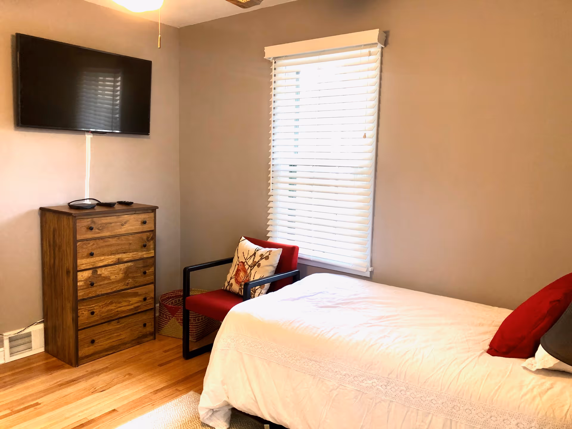 A small bedroom with a single bed covered in a white bedspread and a red pillow. Next to the bed is a red armchair with a decorative pillow featuring a bird design. A wooden chest of drawers is against the wall with a flat-screen TV mounted above it. The room has light brown walls, a window with white blinds, and wooden flooring.