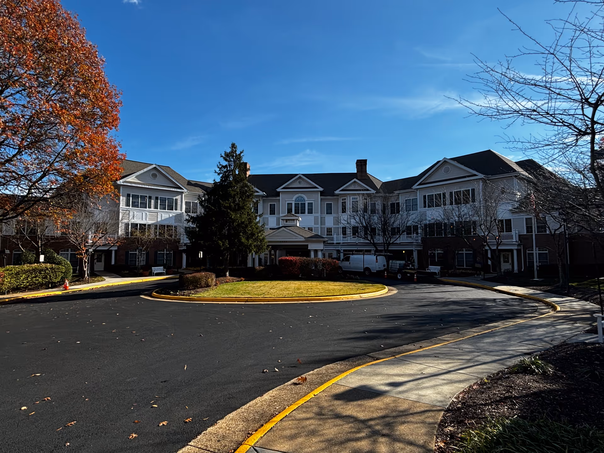 Front exterior of a three-story senior living building with a circular driveway and landscaped grounds under a clear blue sky.