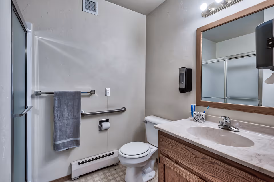 A bathroom with a toilet, a sink with a marble countertop, a large wooden framed mirror, a towel hanging on a metal bar, a soap dispenser mounted on the wall, and a shower with frosted glass sliding doors.