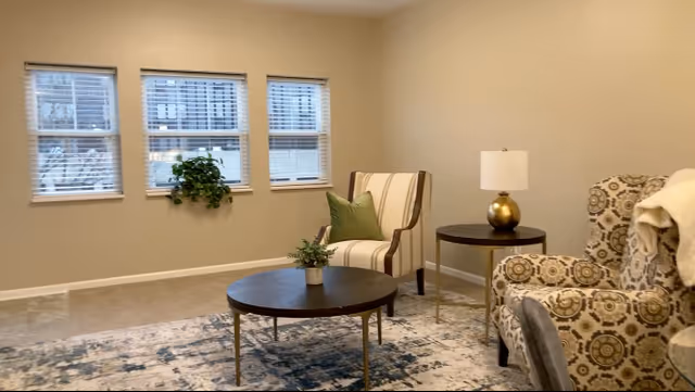 A cozy living room area with three windows on the wall, a hanging green plant, a striped armchair with a green pillow, a round wooden coffee table with a small plant, a patterned armchair with a white throw blanket, and a round side table with a lamp.