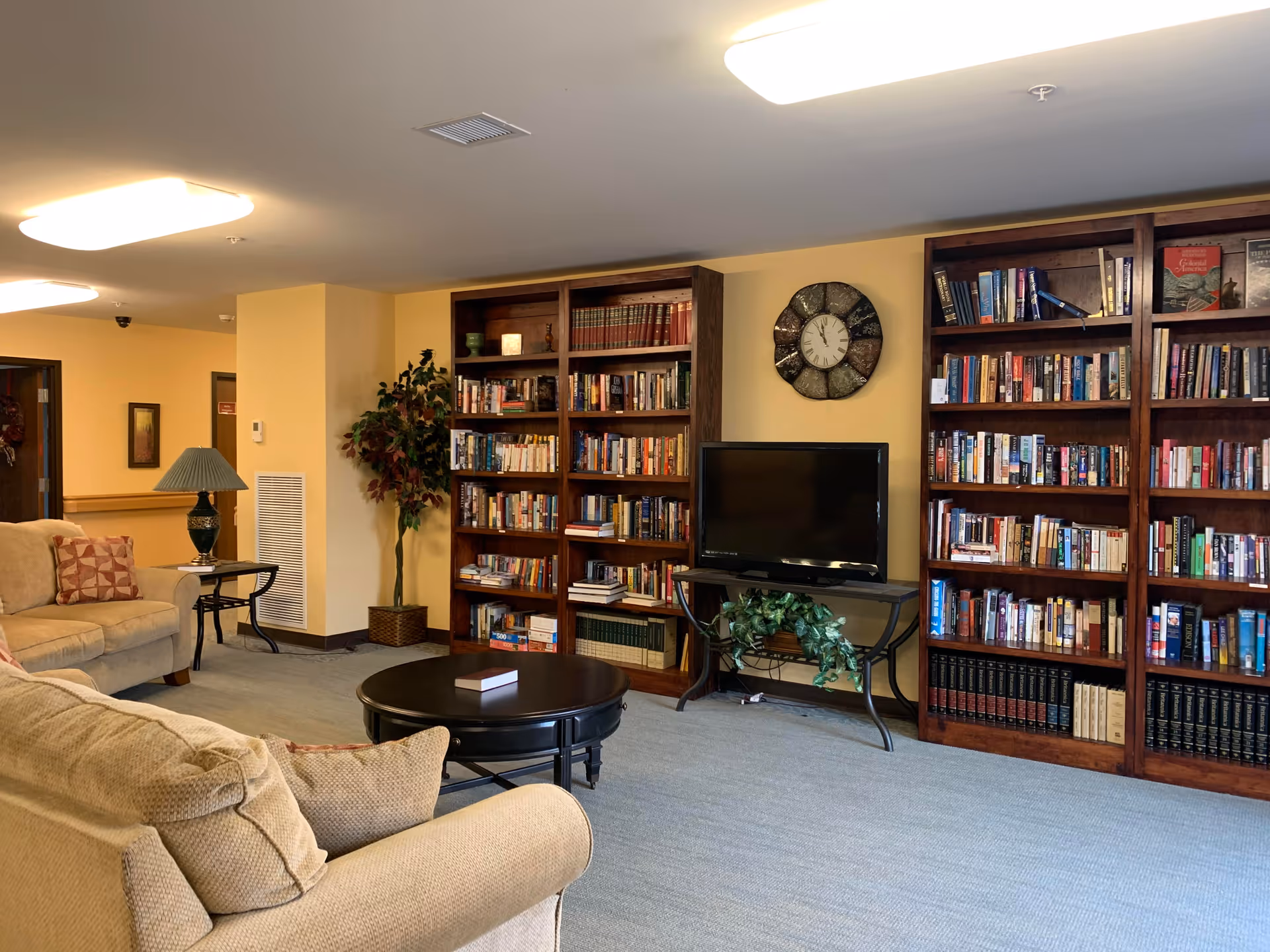 A cozy living room area with beige sofas, a round black coffee table, a table lamp on a side table, and large wooden bookshelves filled with books. A flat-screen TV is placed on a stand between the bookshelves, and a decorative wall clock hangs above the TV. The room has soft yellow walls and carpeted flooring.