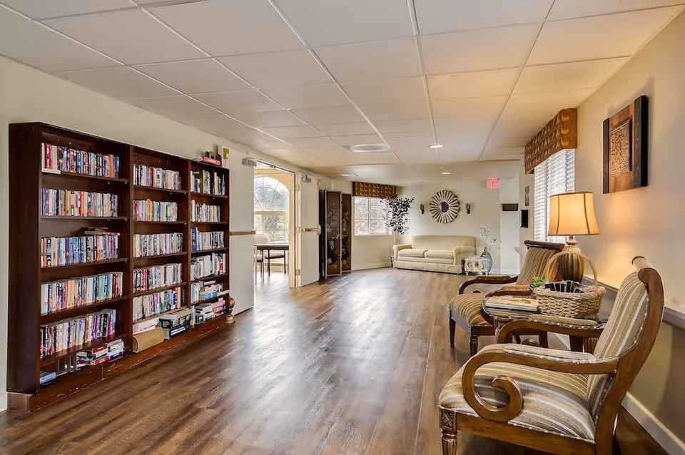 A quiet common lounge with bookshelves along the left wall, wooden chairs and a side table on the right, and a sofa at the far end.