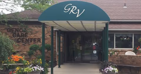 Entrance to Genoa Care Center with a green awning displaying the initials 'GRV'. The building is made of brick with windows and flower beds on either side of the walkway leading to the glass doors.
