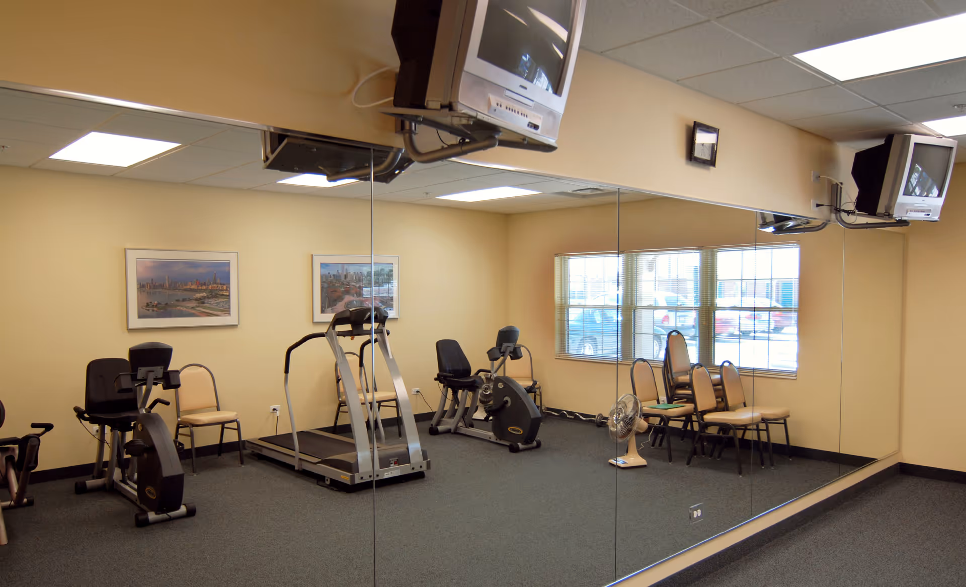 A small exercise room with a treadmill, stationary bikes, several chairs, a wall-mounted TV, and large mirrors covering one wall. The room has beige walls and a window showing parked cars outside.