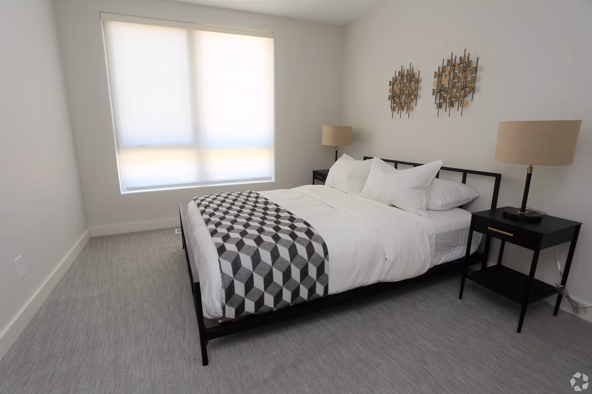 Minimalist bedroom with a made bed featuring white bedding and a geometric throw, two nightstands with lamps, and a large window with blinds.
