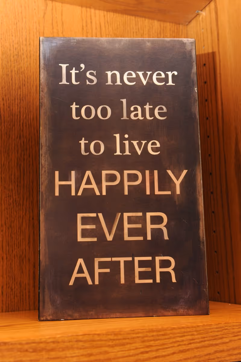 A wooden decorative sign on a shelf that reads "It's never too late to live HAPPILY EVER AFTER."