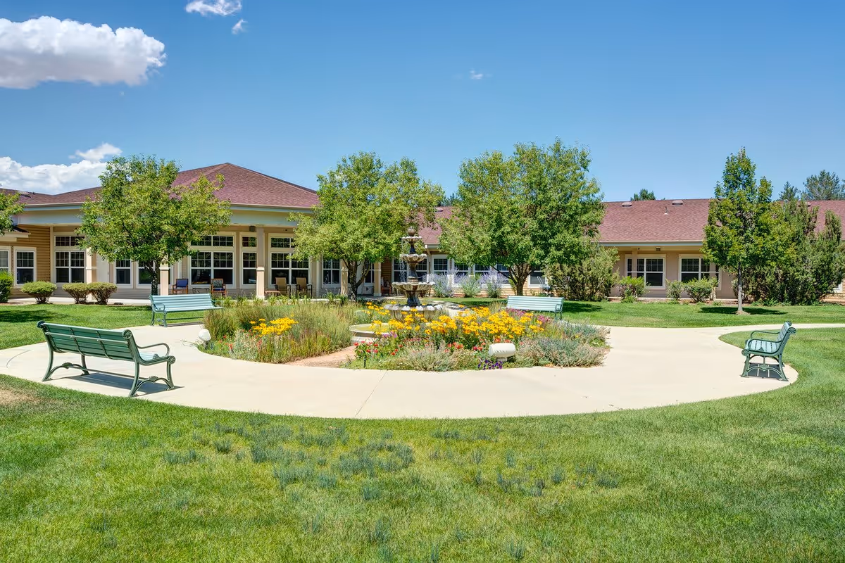 Outdoor garden area at The Courtyards at Mountain View featuring a circular walkway surrounding a flower bed with yellow flowers and a multi-tiered fountain in the center. Several green benches are placed around the walkway, and trees and shrubs are visible near the building with a red roof under a clear blue sky.