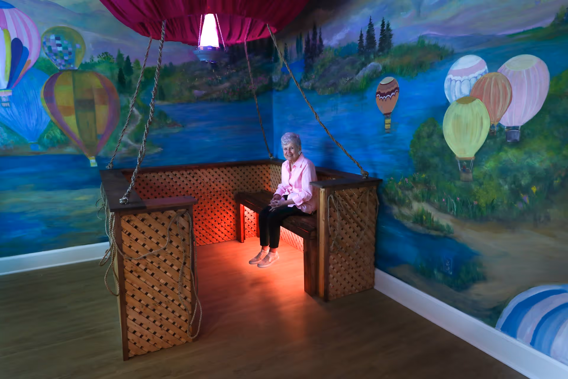 An elderly woman sitting on a wooden swing bench indoors, surrounded by colorful murals of hot air balloons and a scenic landscape on the walls.