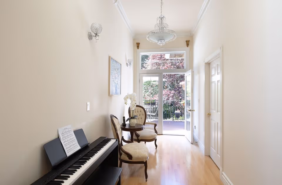 Bright narrow sitting area with a digital piano, two upholstered chairs and a small table leading to glass doors opening onto a deck.
