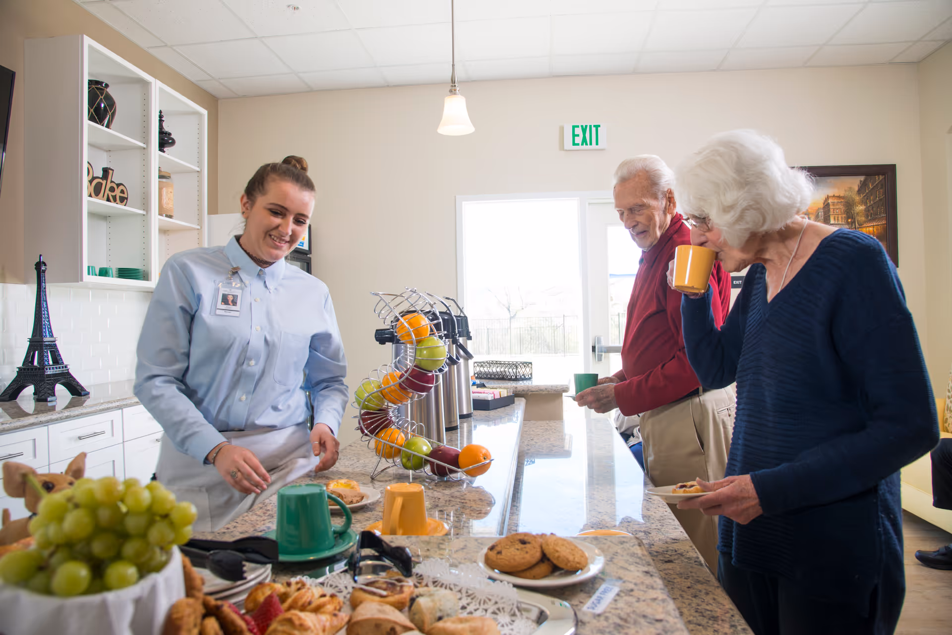 A young female staff member in a light blue shirt serves snacks to two elderly residents, a man and a woman, who are enjoying drinks and pastries at a kitchen counter in a senior living facility. The counter has a fruit holder with apples and oranges, coffee dispensers, and plates of cookies and pastries. The room is bright with a door and exit sign in the background.