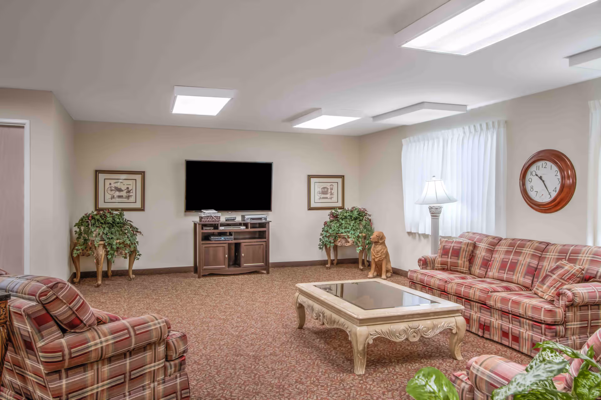 A cozy living room with plaid upholstered armchairs and a sofa arranged around a decorative coffee table with a glass top. The room has beige walls, a carpeted floor, a flat-screen TV mounted on the wall above a wooden TV stand, two framed pictures, two potted plants on wooden stands, a floor lamp, a wall clock, and a decorative dog statue near the window with white curtains.