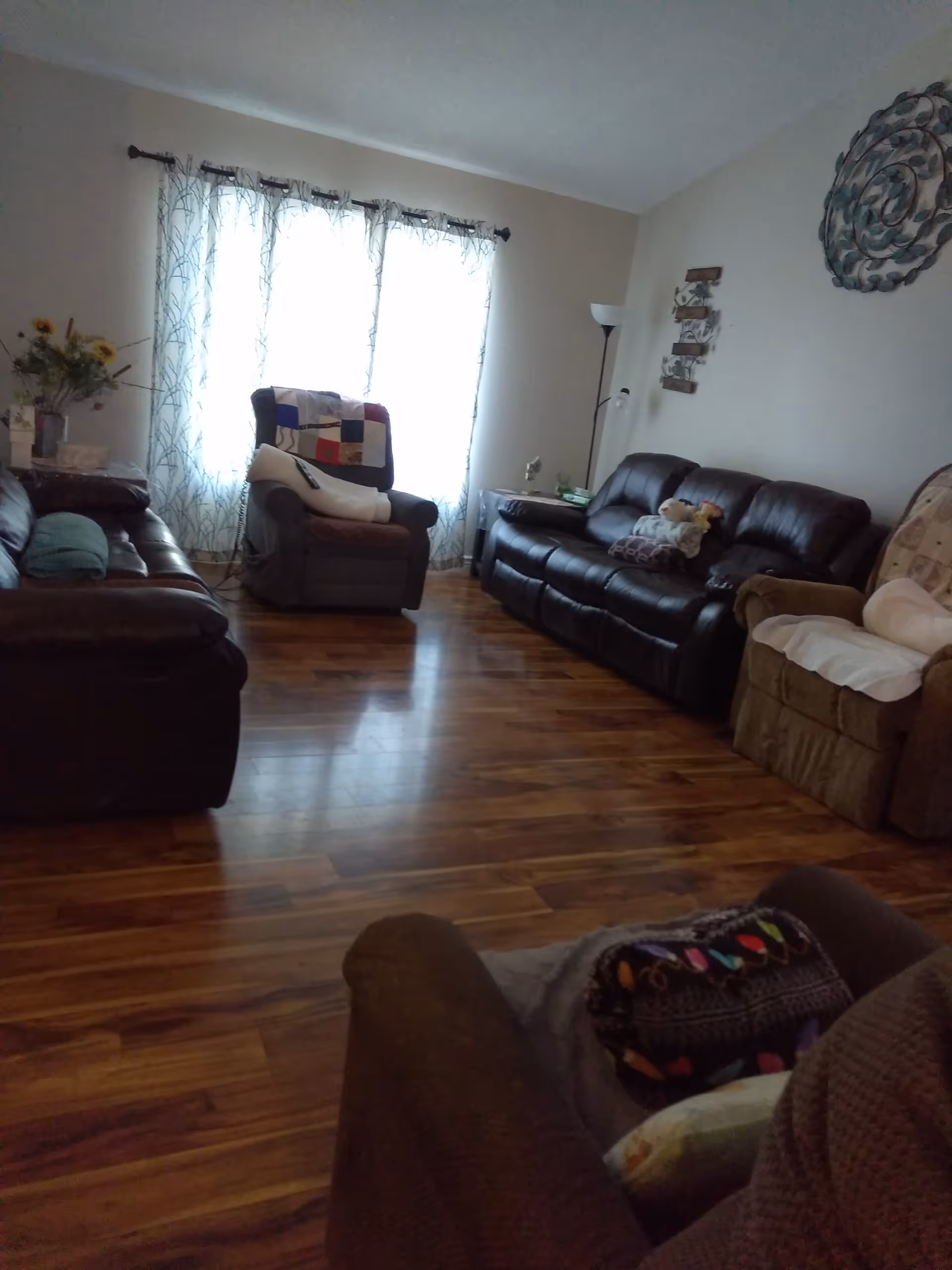 A cozy living room with wooden flooring, featuring multiple armchairs and sofas arranged around the room. A large window with sheer curtains allows natural light to fill the space. Decorative wall art and a floor lamp are visible, along with some cushions and a quilt on the chairs.