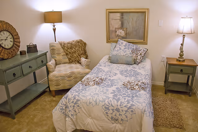 A cozy bedroom with a single bed covered in a floral patterned comforter and matching pillows. Next to the bed is a small wooden nightstand with a lamp. Across from the bed is a cushioned armchair with a throw blanket draped over it. A green console table with a large decorative clock and small items is against the wall. A framed artwork hangs above the bed, and the room is softly lit by two lamps.