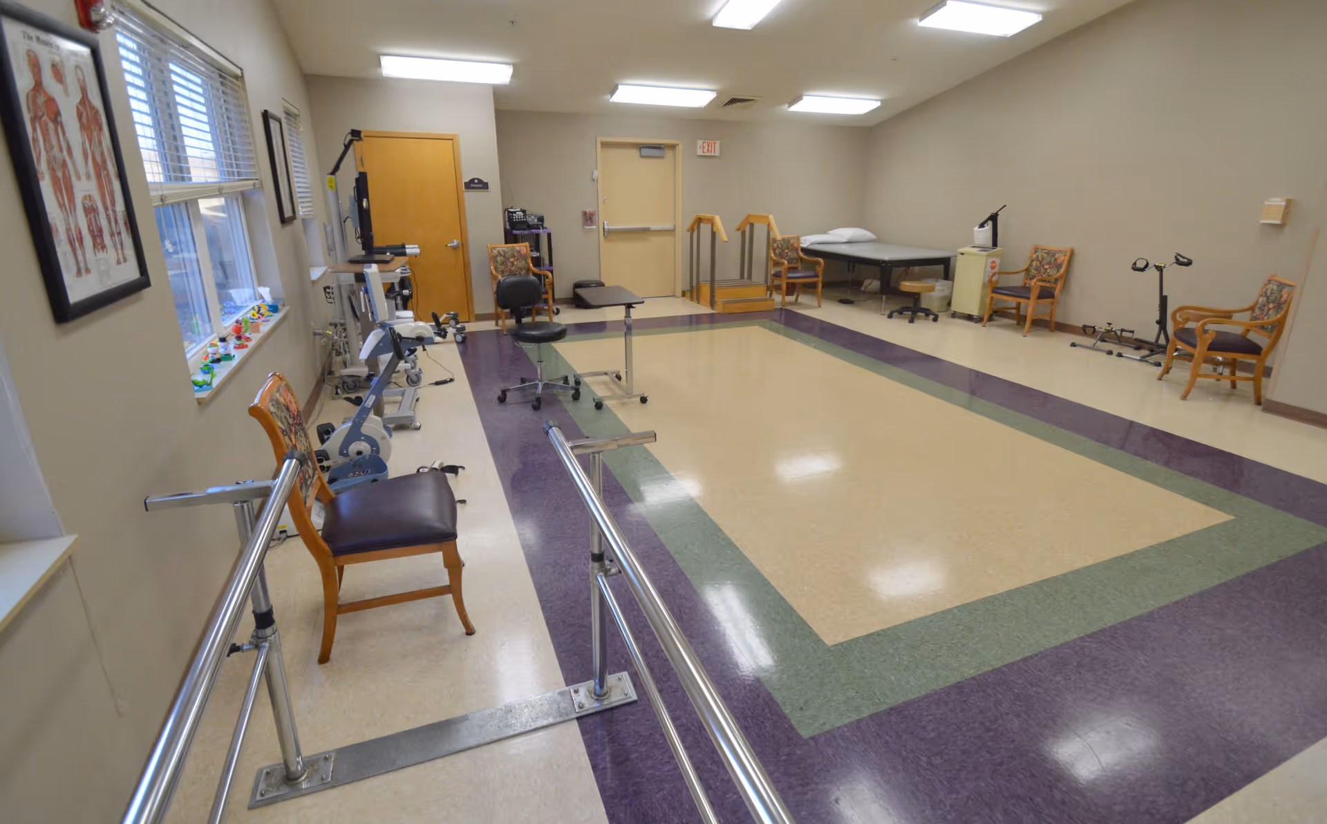 A spacious physical therapy room with parallel bars for walking exercises, several chairs with floral cushions, exercise equipment including a stationary bike, a treatment table, and anatomical charts on the wall. The room has large windows letting in natural light and a door at the far end.
