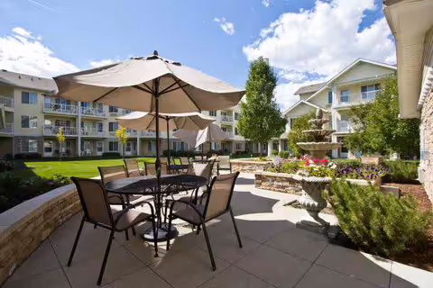 Sunlit courtyard patio with tables and umbrellas, a fountain, and multi-story retirement community buildings surrounding a landscaped lawn.