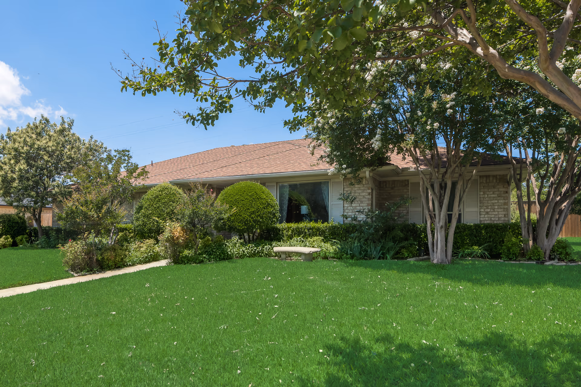 Single-story brick building with a brown shingled roof surrounded by well-maintained green lawn, bushes, and trees under a clear blue sky.