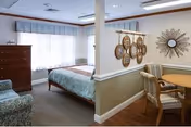 Sunny studio-style room with a bed, dresser, upholstered chairs and a small dining table separated by a half wall.
